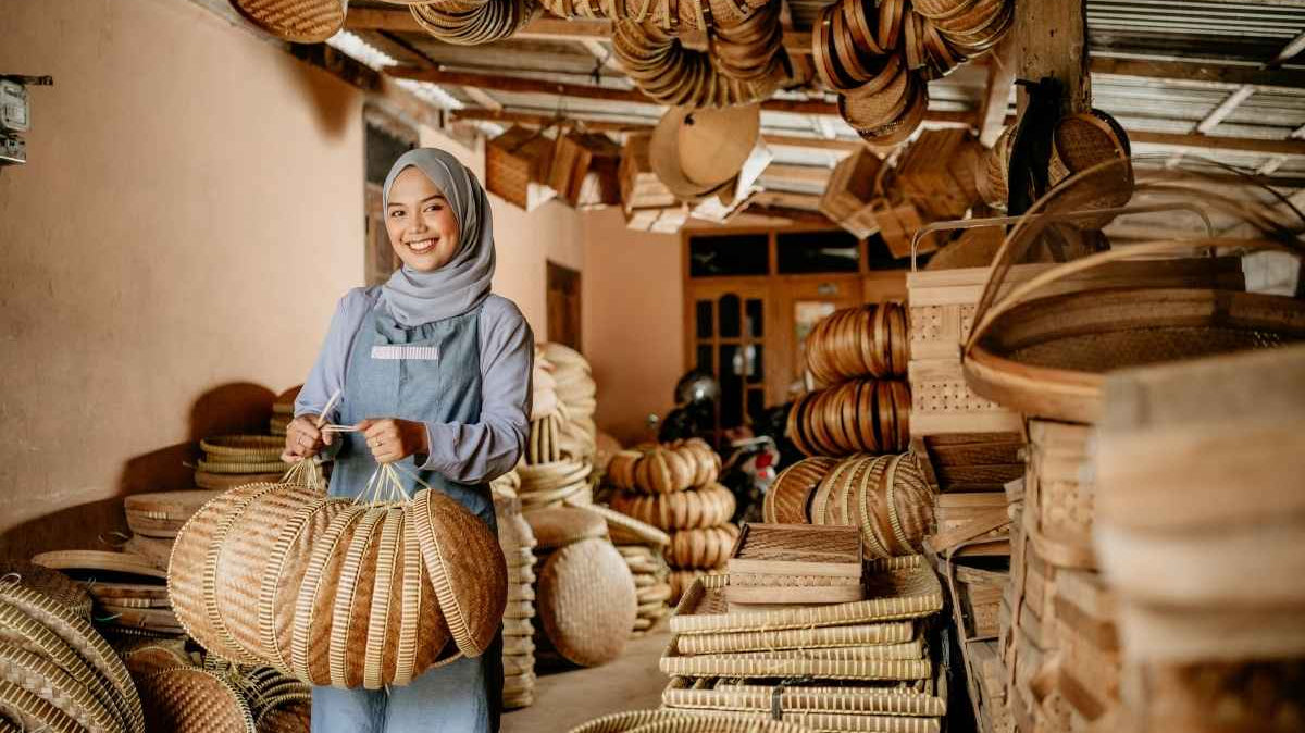 A rural artisan holding several of her handmade crafts, and surrounded by all her other handmade woven crafts in a workshop setting