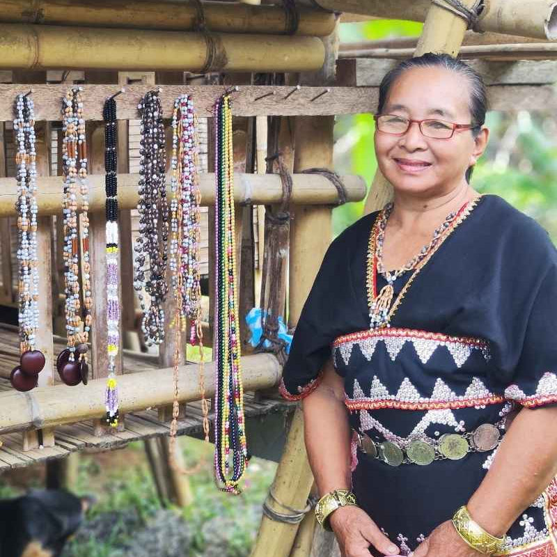Woman in traditional attire standing next to all the handmade beaded necklaces in a natural setting/