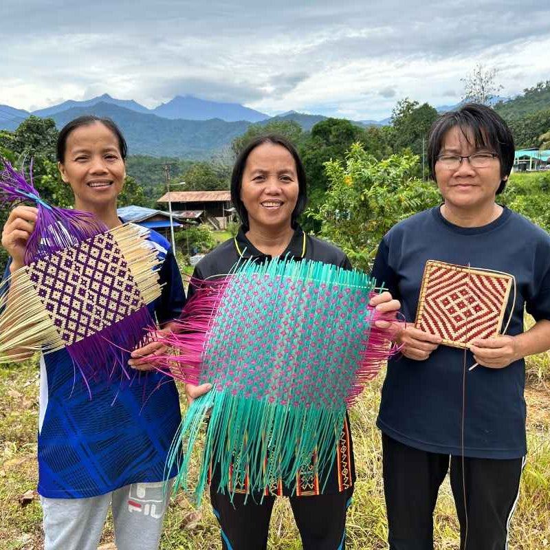 Three people holding handcrafted woven crafts against a natural background with mountains.