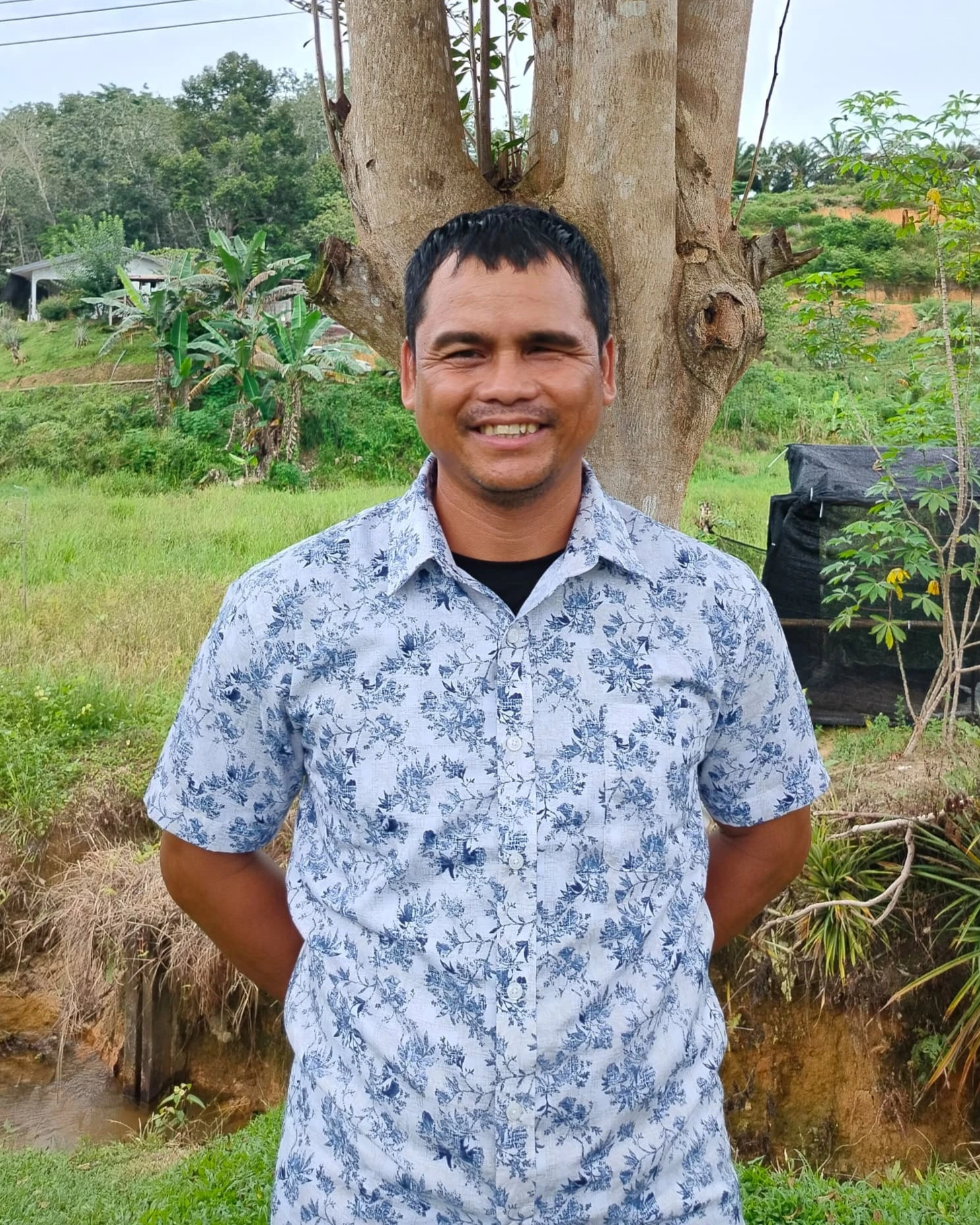 Man standing outdoors in a natural setting with trees and greenery.