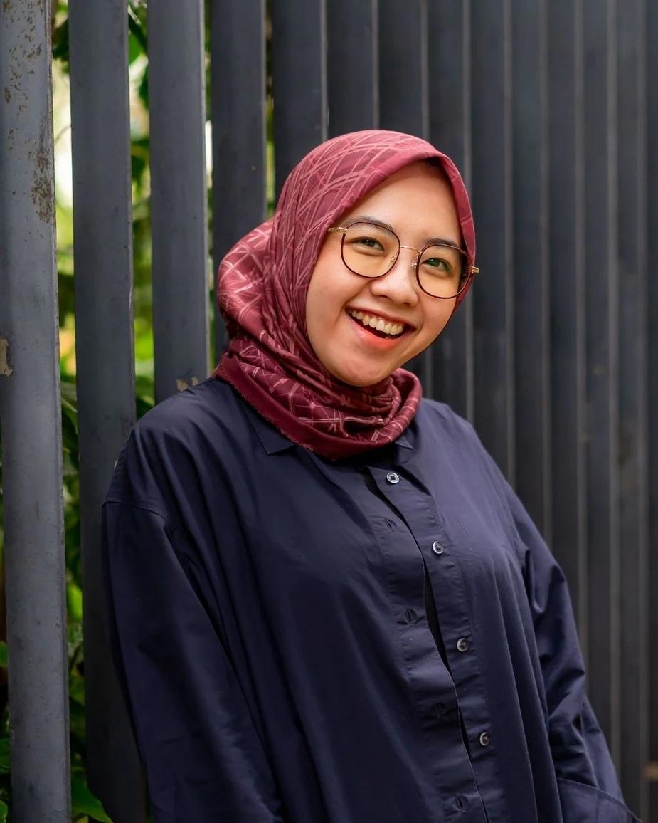 Malay woman wearing a dark blue shirt and pink hijab standing in front of a metal fence.