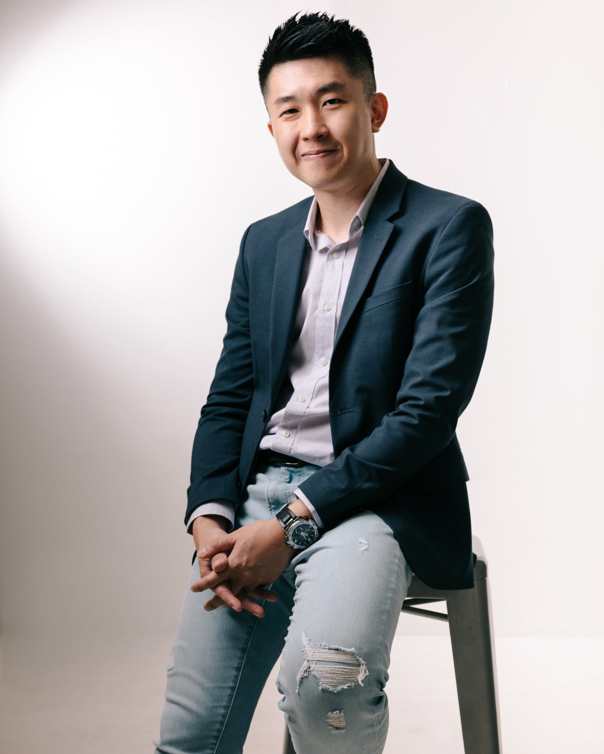 Man in a dark blazer and light jeans sitting on a stool against a plain background