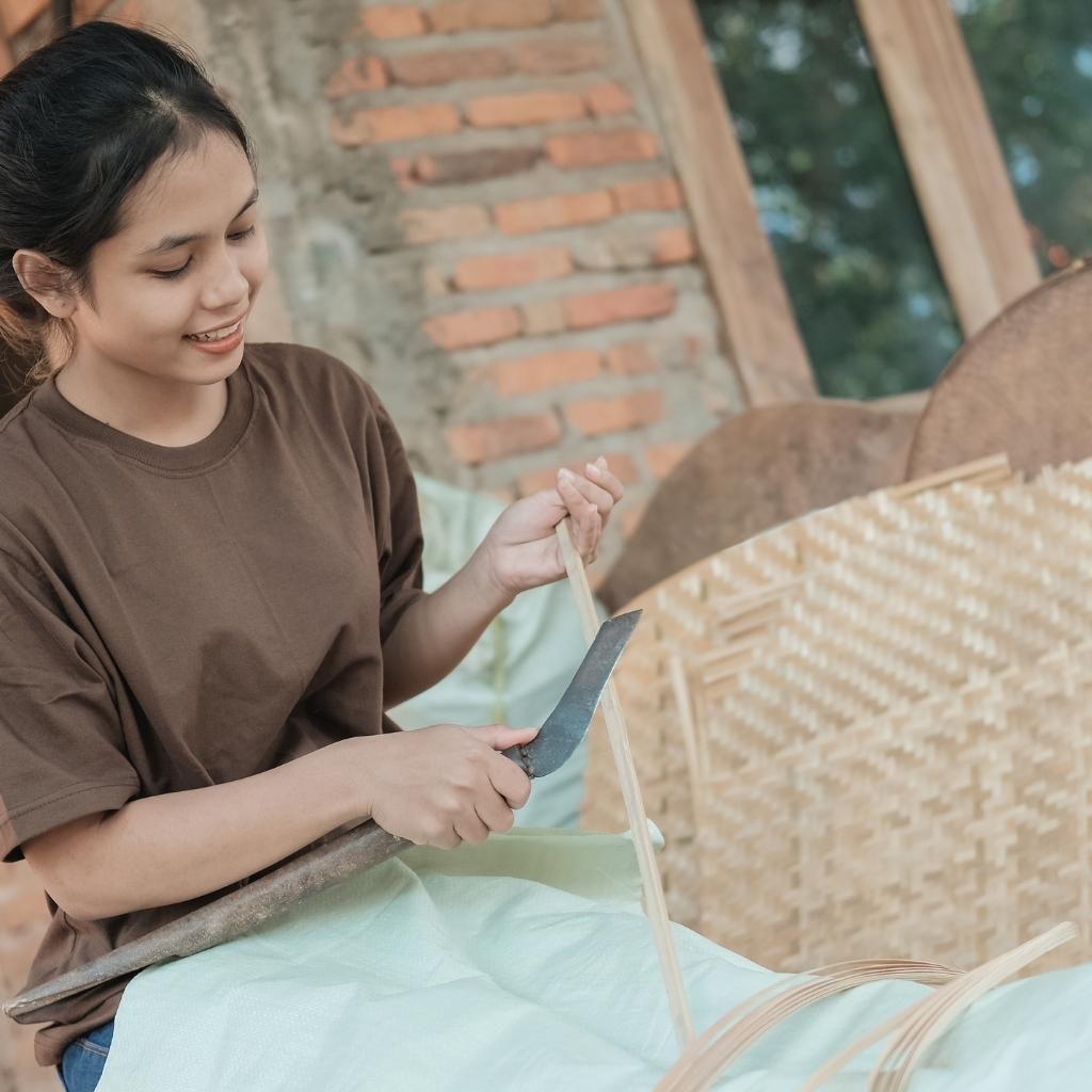 Woman working handcrafting rattan products outdoors, surrounded by natural elements.
