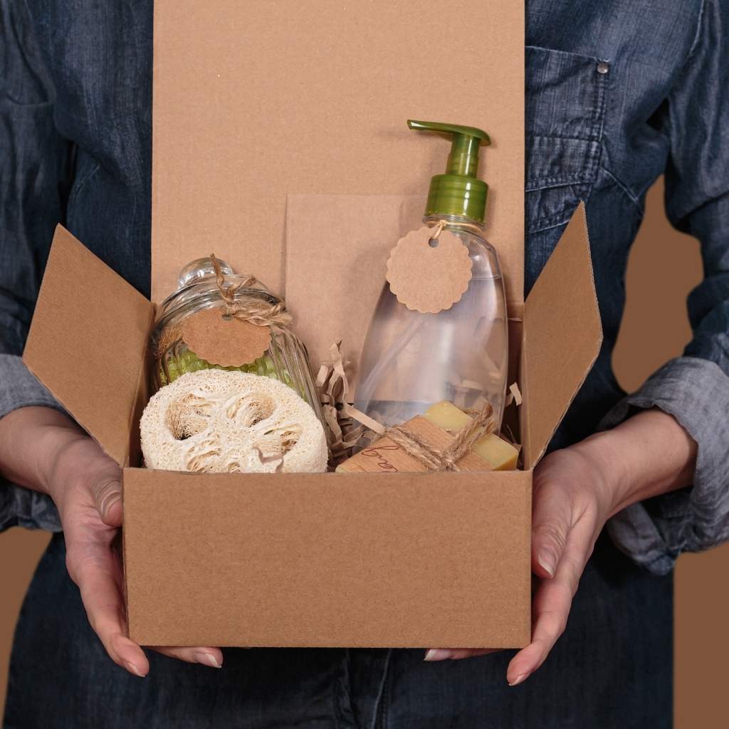 Person holding a cardboard box with eco-friendly products including a bottle, jar, and sponge.