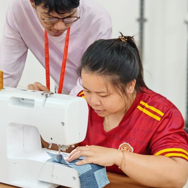 Designer guiding a rural woman on proper sewing techniques using a sewing machine during a hands-on artisan craftsmanship workshop in a classroom setting