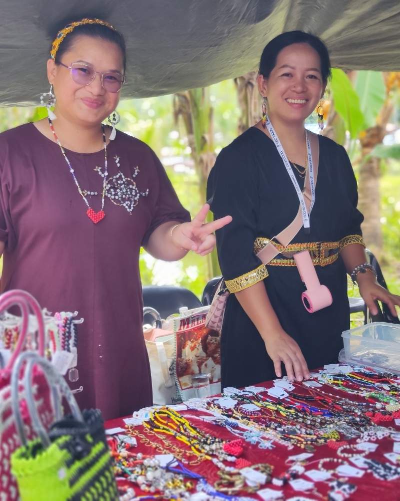 Two women standing behind a table with various handmade crafts, including jewelry, in an outdoor setting.