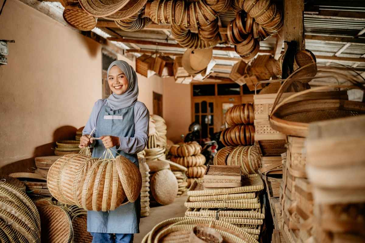A rural artisan holding several of her handmade crafts, and surrounded by all her other handmade woven crafts in a workshop setting