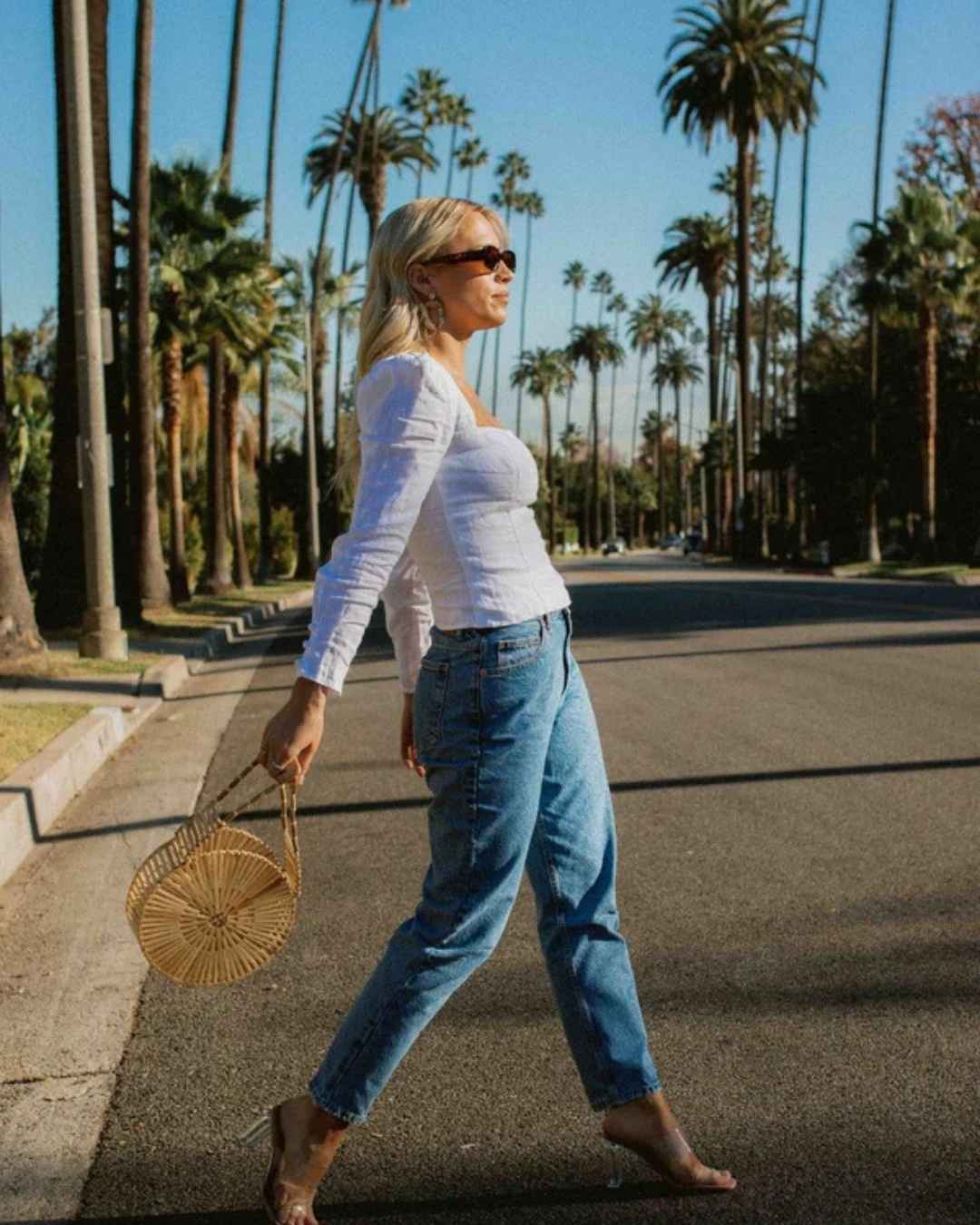 Woman walking on a street lined with palm trees, while holding a rattan bag