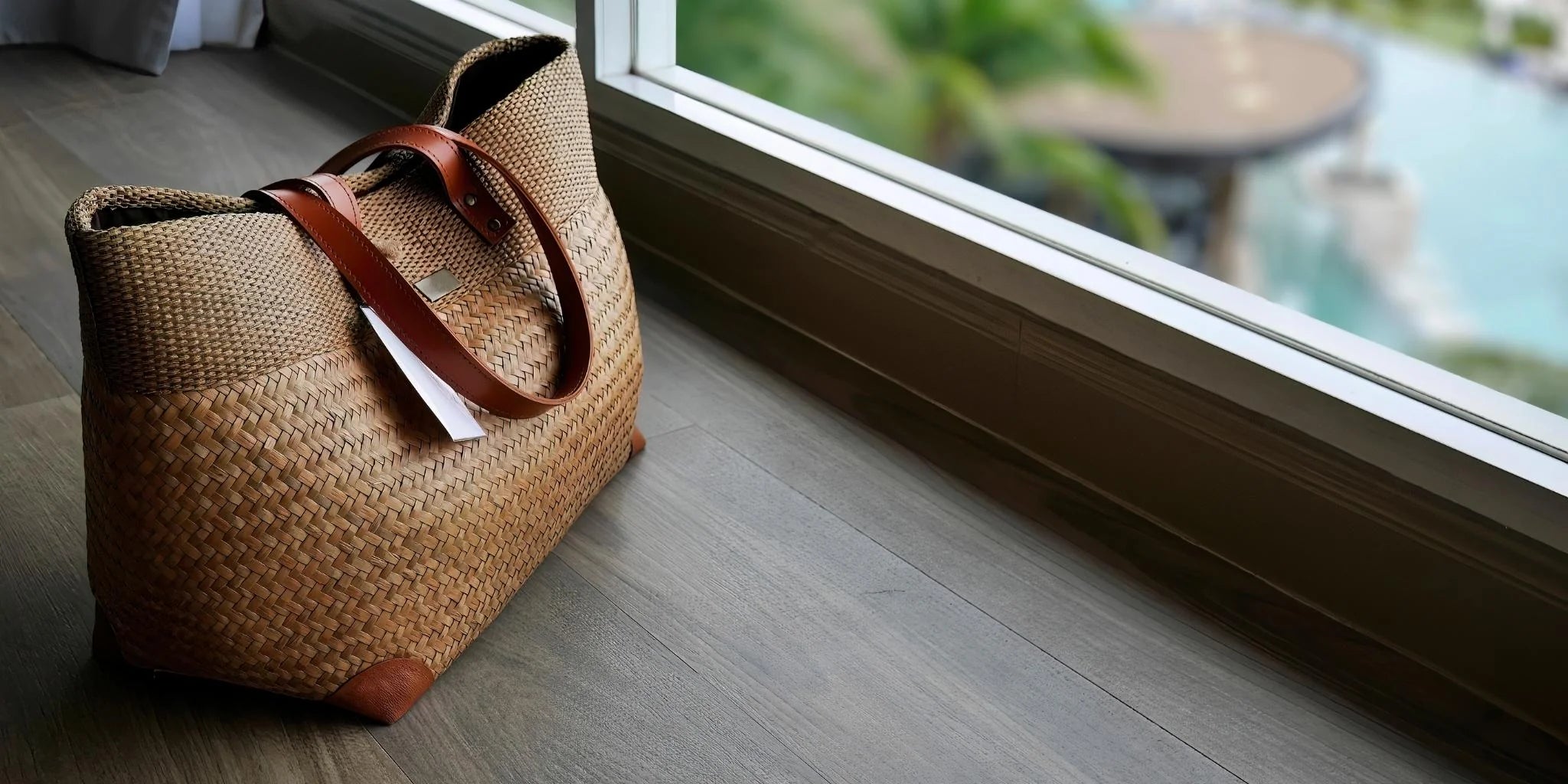 Woven bag with leather handle on a wooden floor near a window with a view of a pool.