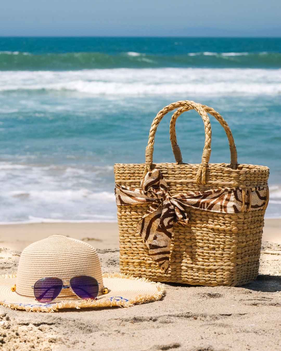 Woven bag with a decorative bow and straw hat on a sandy beach with ocean in the background