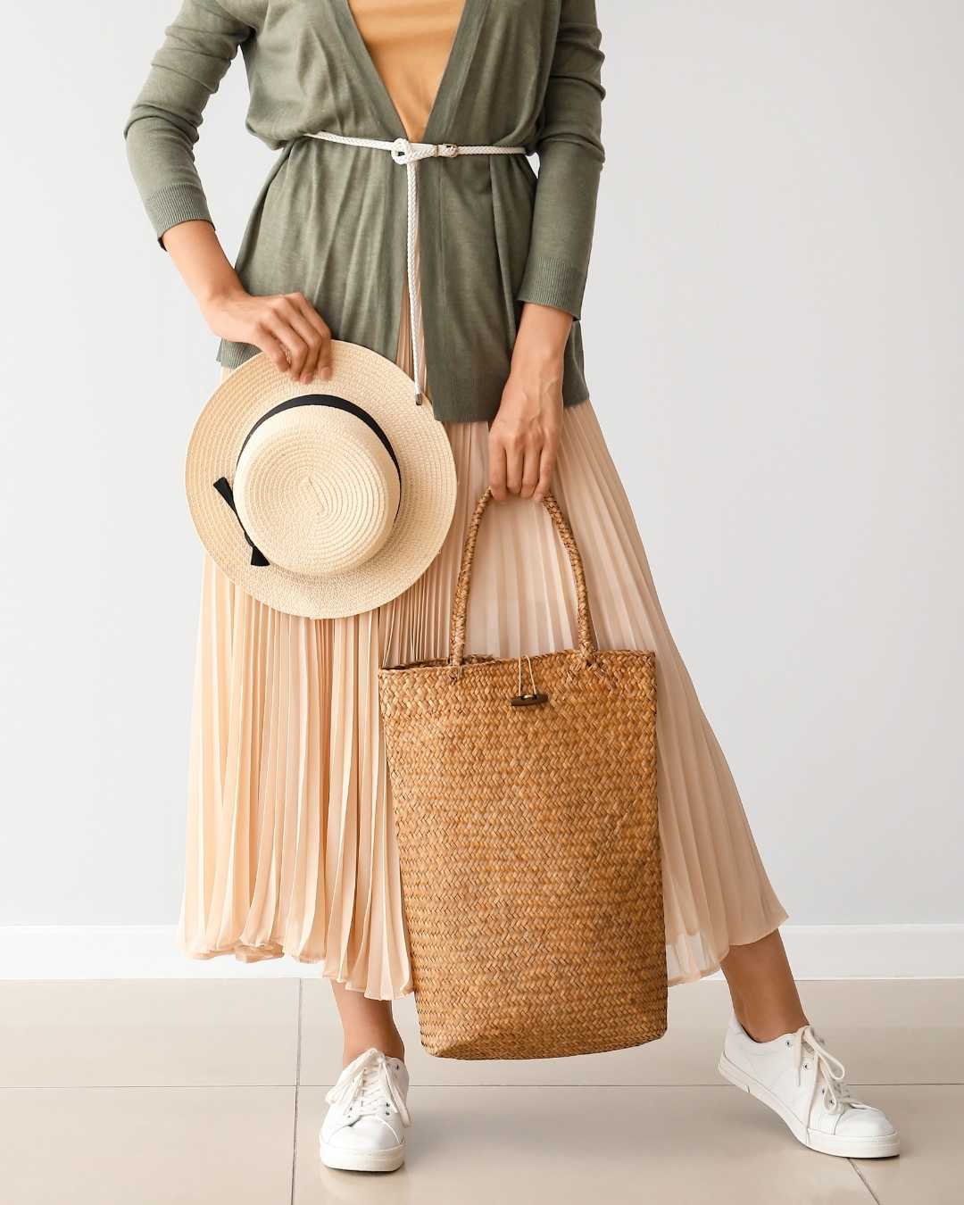 Person holding a straw bag and a hat, wearing a green jacket and beige pleated skirt.