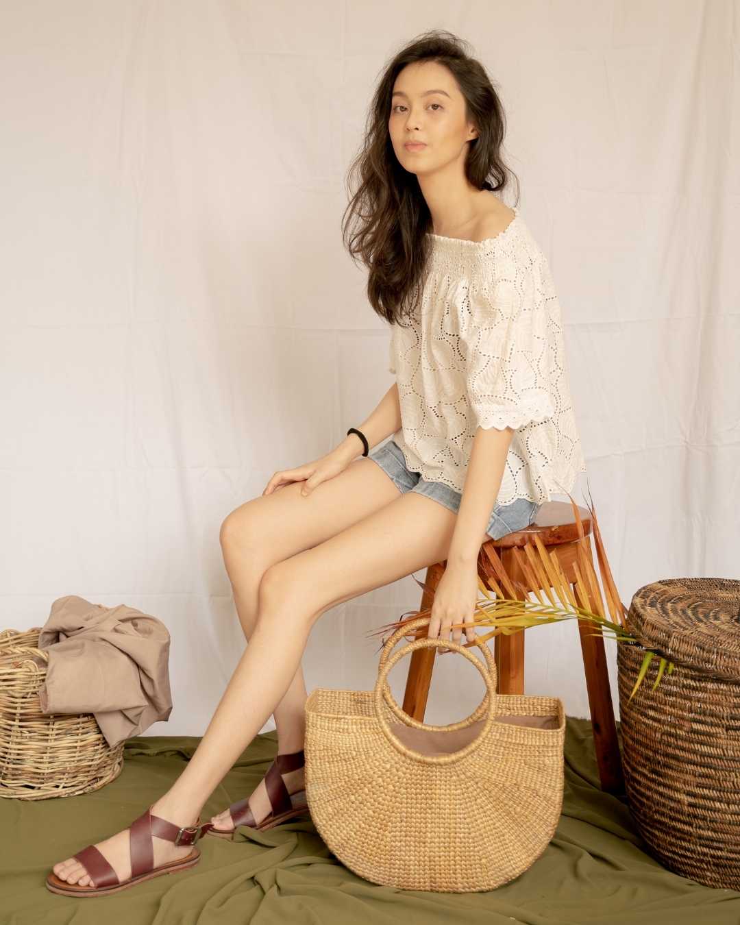 Woman sitting on a wooden stool holding a woven bag, wearing a white lace top and denim shorts.