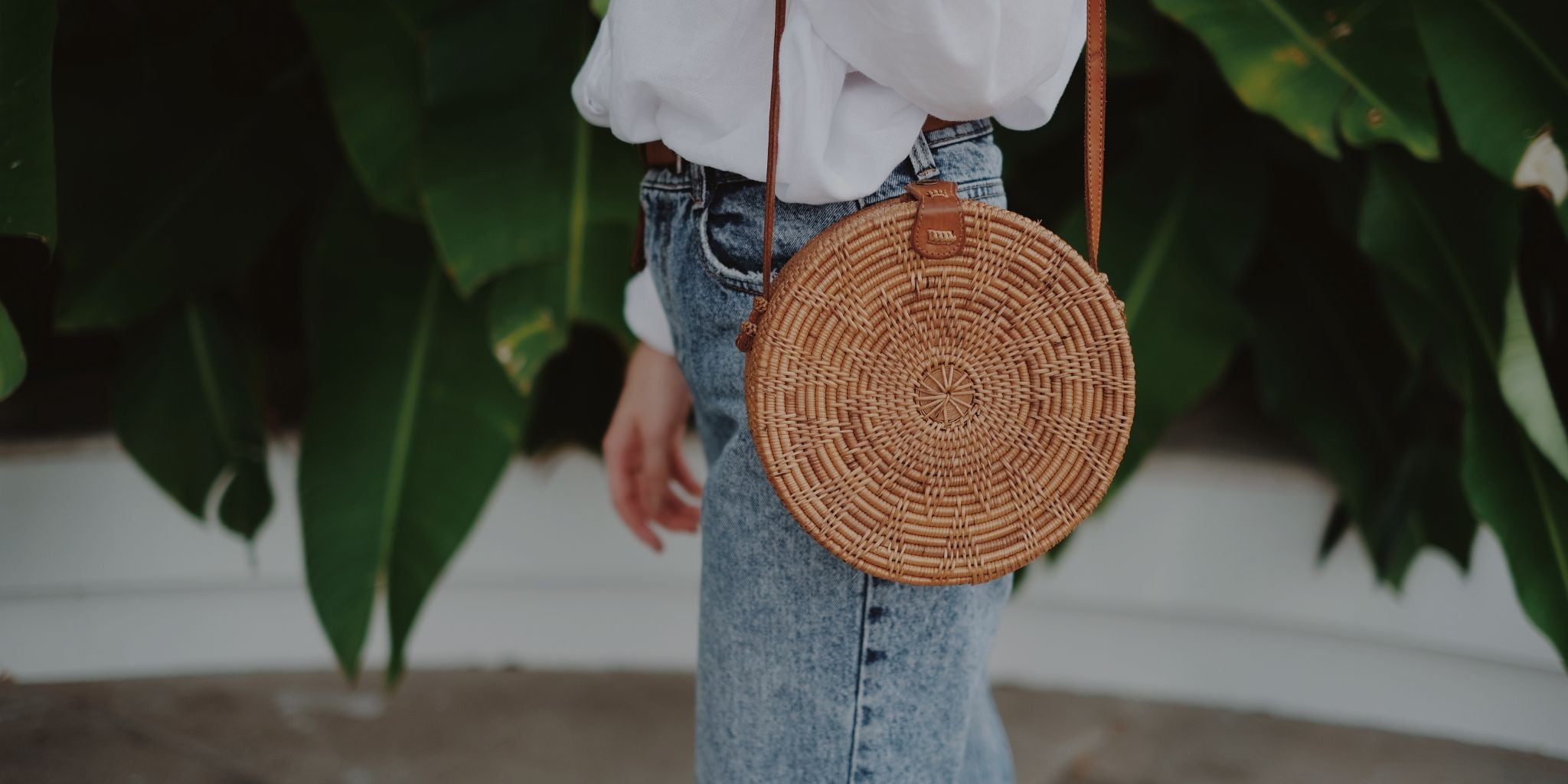 Person wearing a white blouse and blue jeans with a round woven bag over one shoulder, standing against a blurred green leafy background.