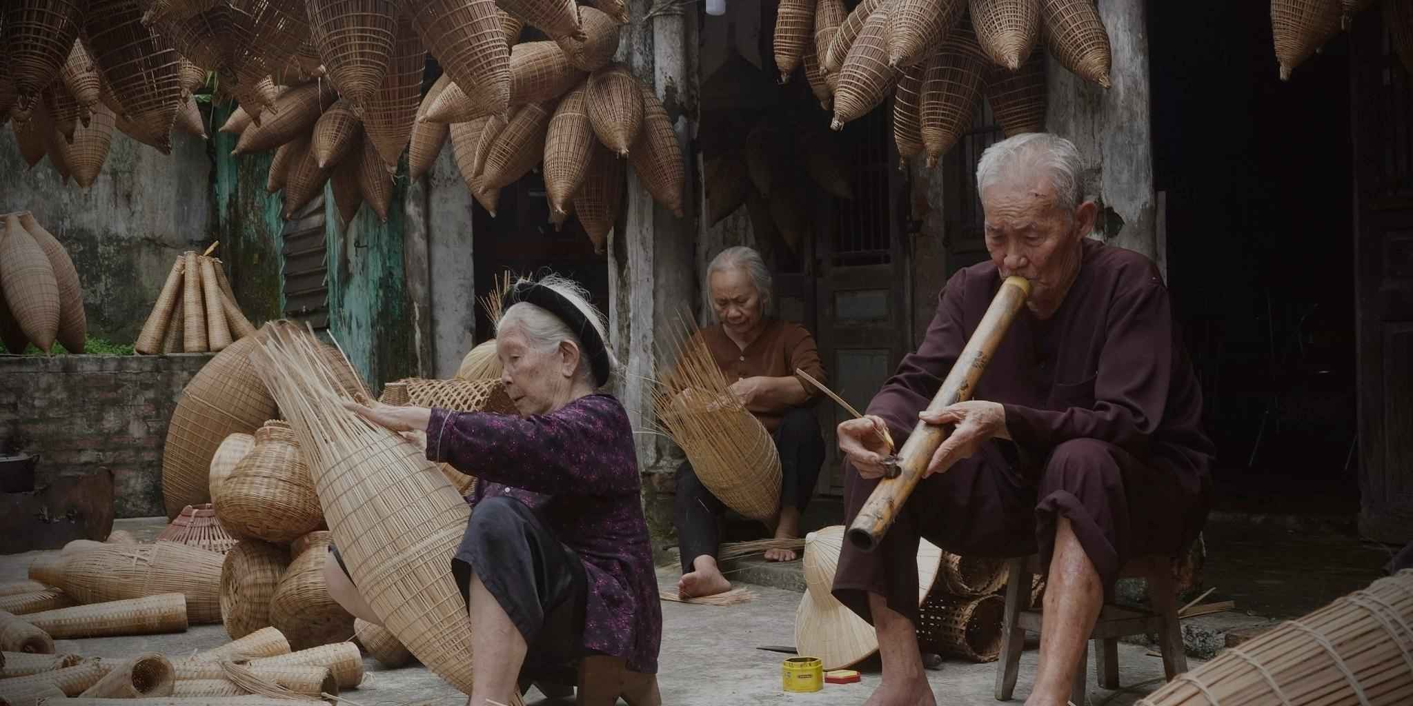 Three elderly individuals working with woven materials in a rustic setting.