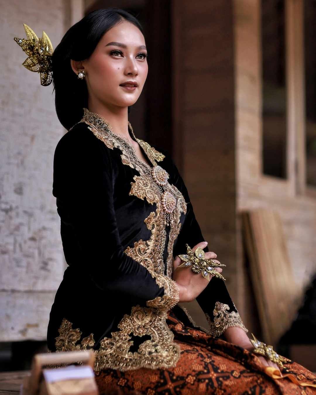 Woman in traditional black and gold outfit with intricate patterns, sitting on a wooden chair.