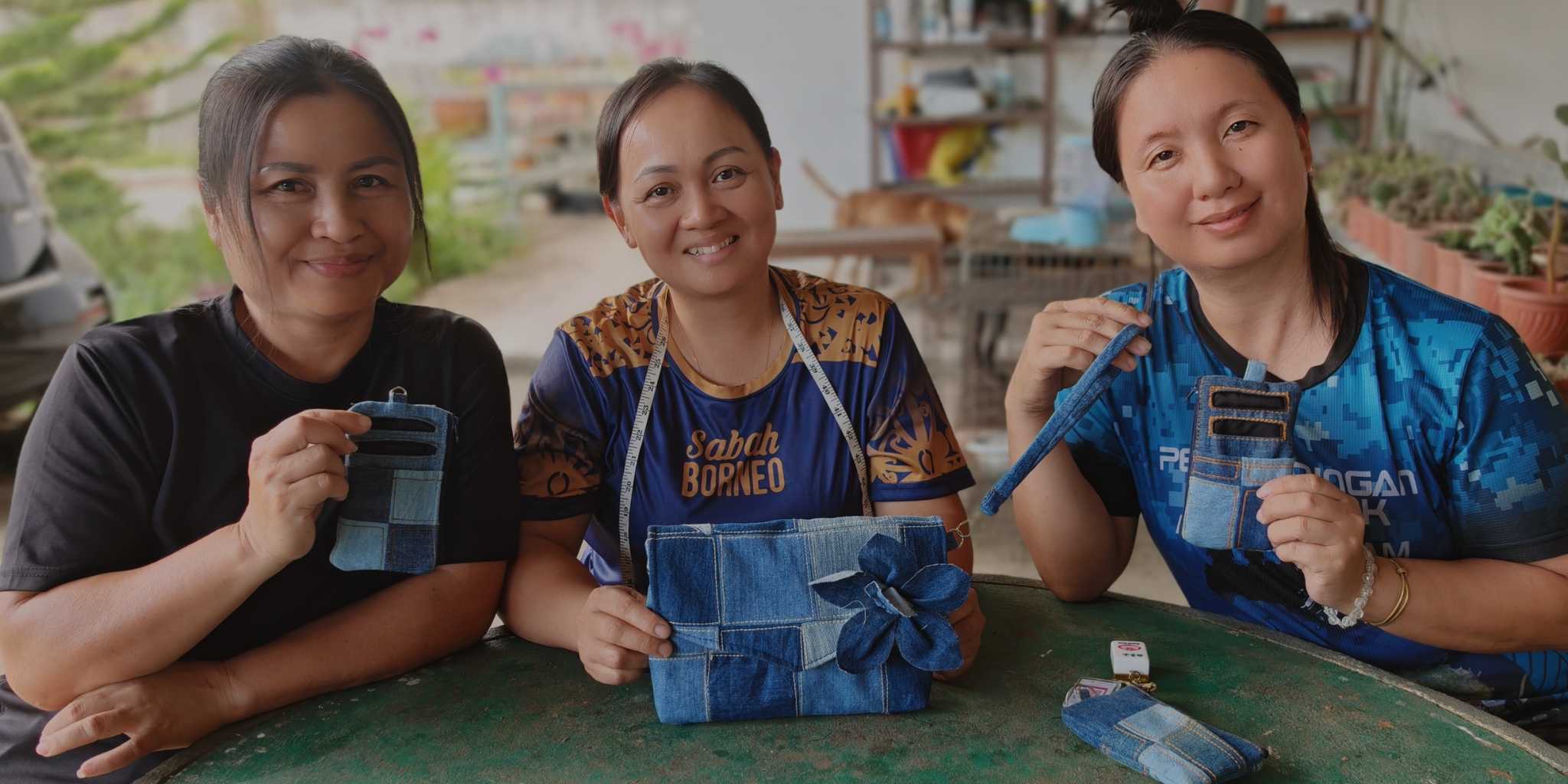 Three women sitting together, each holding an upcycled denim handmade product, in a workshop setting.
