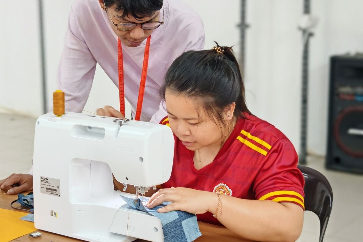 Designer guiding a rural woman on proper sewing techniques using a sewing machine during a hands-on artisan craftsmanship workshop in a classroom setting